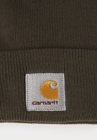 Olivově zelená pletená čepice s tkanou etiketou Carhartt se žlutým emblémem a červeným textem, texturovaná žebrovanými vzory.