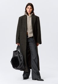 Long dark green coat with double-breasted design, gray knit hoodie underneath, black flared pants, and a textured black bag with fur detail.