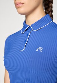 Polo shirt bleu à côtes avec des détails blancs, doté d'un col et de trois boutons, et d'un petit logo brodé sur le côté gauche.