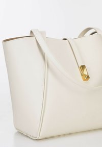 Crème leren tote handtas met dubbele handvaten en een gouden rechthoekige sluiting detail op een handvat, tegen een witte achtergrond.