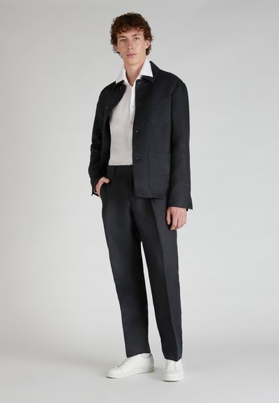 Chaqueta de lino azul marino con cierre de botones, dos bolsillos frontales y una camisa de algodón blanca debajo. Combinada con pantalones oscuros y zapatillas blancas.