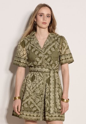 Jeune femme aux longs cheveux blonds portant une robe courte à manches courtes à motif vert olive et des boucles d'oreilles créoles, un bracelet et un jonc en or.