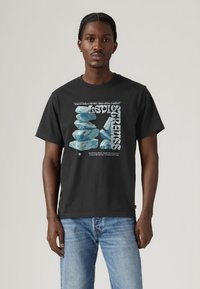 Μαύρο βαμβακερό T-shirt με γραφικό σχέδιο που περιλαμβάνει στοιχεία που μοιάζουν με μπλε πέτρες και λευκό κείμενο, συνδυασμένο με τζιν τζιν σε ανοιχτή απόχρωση.