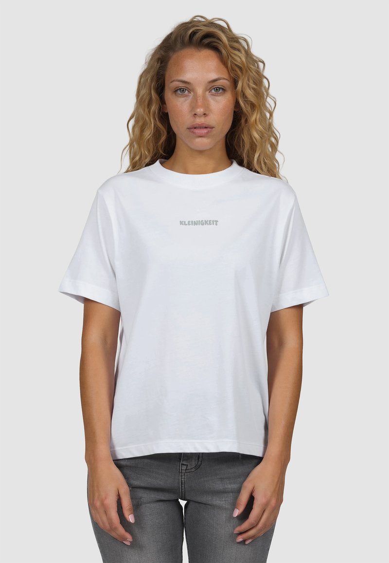 Femme aux cheveux bouclés portant un t-shirt blanc avec le texte "KLEINIGKEIT" et un jean gris, debout devant un fond gris clair.