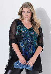Schwarzes, transparentes Oberteil mit lebhaftem Pfauenfederdesign in Blau- und Grüntönen. Getragen mit einer blauen strukturierten Clutch und schwarzen Leggings.