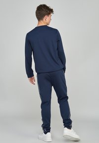 Camisa de manga larga azul marino combinada con pantalones de chándal azul marino que presentan franjas laterales blancas y zapatillas blancas, enfatizando un estilo atlético relajado.