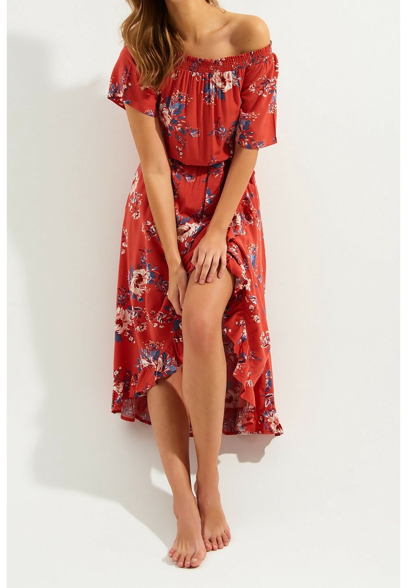 Femme portant une robe fleurie rouge à épaules dénudées avec un ourlet à volants, tenant légèrement la robe soulevée pour dévoiler ses jambes, debout pieds nus.
