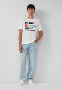 Junger Mann in einem weißen T-Shirt mit dem Regenbogen-Aufdruck "Human Beings 100% Organic Colors may vary", hellblauen Jeans und weißen Turnschuhen.