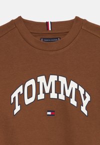 Sweatshirt marron avec "TOMMY" en grandes lettres blanches bordées de bleu marine et un petit emblème en rouge, blanc et bleu en bas.