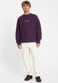 Sweatshirt violet avec le logo "VOLCOM" sur la poitrine, associé à un pantalon blanc et des chaussures noires avec des accents jaunes. Design simple et décontracté.