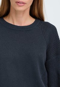 Donker marineblauwe trui met een geribbelde ronde hals, gestructureerde gebreide mouwen en een zachte, gladde body met een lichte glans. Zichtbare naaddetails.