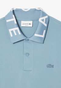 Niebieska slim-fit koszulka polo z dzianym kołnierzykiem, białym napisem, dwoma guzikami przy zapięciu oraz haftowanym logo krokodyla na klatce piersiowej.