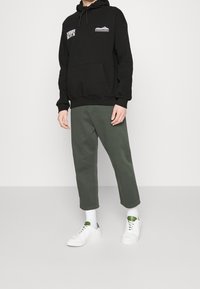 Felpa nera con cappuccio e stampe, abbinata a pantaloni verdi oliva. Scarpe da ginnastica bianche con accenti verdi e calzini bianchi. Stile minimalista.