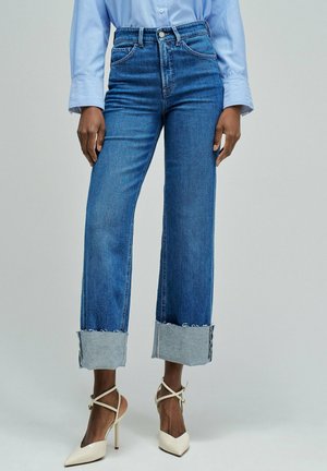 Pantalones vaqueros de mezclilla azul de tiro alto con piernas anchas y un puño deshilachado azul claro. Combinados con zapatos de tacón alto de color crema.