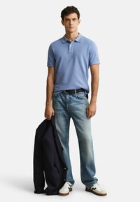 Vaaleansininen polo-paita, farkut ja valkoiset lenkkarit, joissa on mustia yksityiskohtia. Laivastonsininen bleiseri vasemmassa kädessä; rento istuvuus ja sileä lämpö.