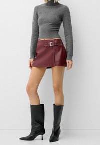 Spódnica z bordowej skóry z bocznym rozporkiem i paskiem zdobionym ćwiekami, zestawiona z dopasowanym szarym golfem i czarnymi kozakiem za kolano z noskiem w szpic.