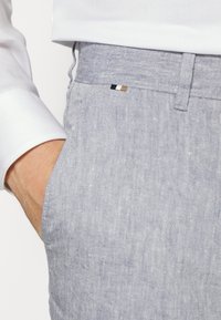 Hand in pocket wearing light blue textured pants and a white long-sleeve shirt with a small black, white, and tan fabric tag on the waistband.