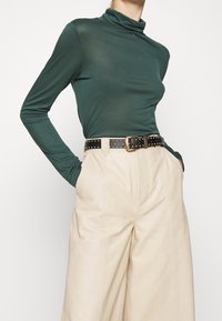 Grünes Langarm-Turtleneck-Shirt, kombiniert mit beigen weiten Hosen. Das Outfit wird durch einen schwarzen Nieten-Gürtel ergänzt, um zusätzliche Details zu verleihen.