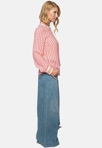 Rot und rosa gemusterte Bluse mit einem weißen Kragen, kombiniert mit hoch sitzenden blauen Jeans. Die Bluse hat gestreifte Manschetten und einen lockeren Schnitt.