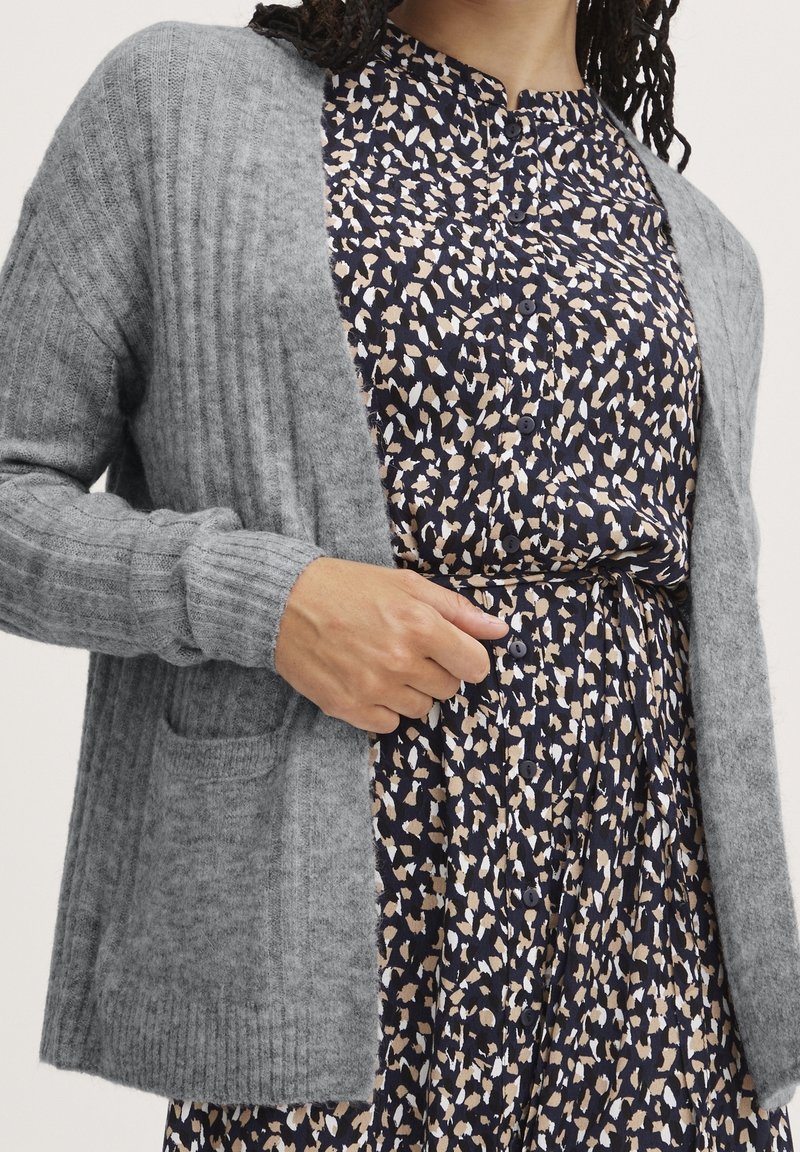 Cardigan gris tricoté avec texture côtelée, doté d'une poche frontale, porté sur une robe bleu marine à motif abstrait beige et blanc avec des boutons.