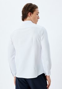 Camisa blanca de manga larga con una tela texturizada, cuello con puntas y puños abotonados. La parte trasera presenta un diseño limpio y ajustado.