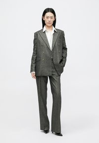 Sivi prugasti dvostruko zatvoren sako u kombinaciji s odgovarajućim hlačama; tkanina izgleda teksturirano; ispod bijela košulja; elegantne, šiljate cipele.