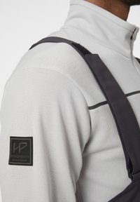 Top gris de cuello alto con tirantes negros, que presenta una tela texturizada y un parche de logo negro en el hombro izquierdo, destacando un diseño elegante.
