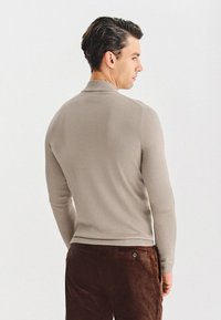 Beżowy dziergany sweter z wysokim kołnierzem, dopasowanymi rękawami i gładką fakturą. Brązowe spodnie ze sztruksu z detalem kieszeni z tyłu.