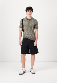 Polo shirt à motifs olive avec des manches courtes, associé à un short noir. Le modèle porte des sneakers blanches et un sac messager noir.