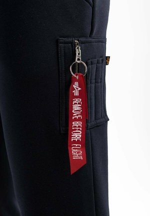 Marineblaue Sweatpants mit einer Seitentasche, ausgestattet mit einem silbernen Reißverschluss und einem roten Etikett, auf dem "VOR FLUG ENTFERNEN" steht. Das Material ist weich und hat eine strukturierte Oberfläche.