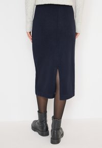 Femme portant une jupe midi bleu marine avec une fente à l'arrière, des collants noirs transparents, des bottes noires à cheville et un pull en maille torsadée gris clair.