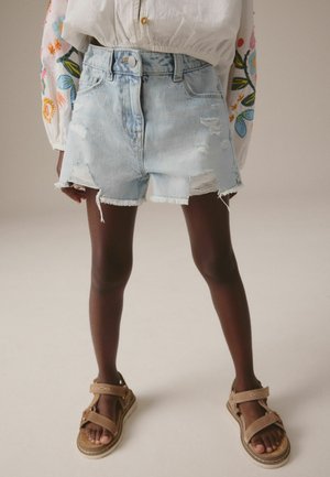 Pantalones cortos de mezclilla azul claro con dobladillos deshilachados y detalles desgastados, combinados con sandalias marrones con correas ajustables.