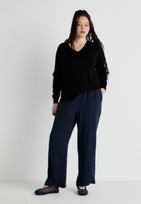 Suéter preto de decote em V combinado com calças largas de riscas azul-marinho, apresentando um corte descontraído. O modelo usa sapatos casuais com detalhes brancos.