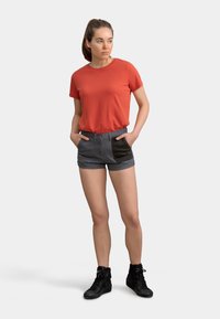 T-shirt rouge à manches courtes, shorts gris avec des poches à rabats noires et chaussures montantes noires. Le modèle se tient avec les mains dans les poches, contre un fond neutre.