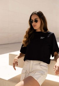 Zwart cropped t-shirt met logo, gecombineerd met lichtbeige cargoshorts met patch pockets en contrasterende stiksels. Vierkante zonnebril gedragen.