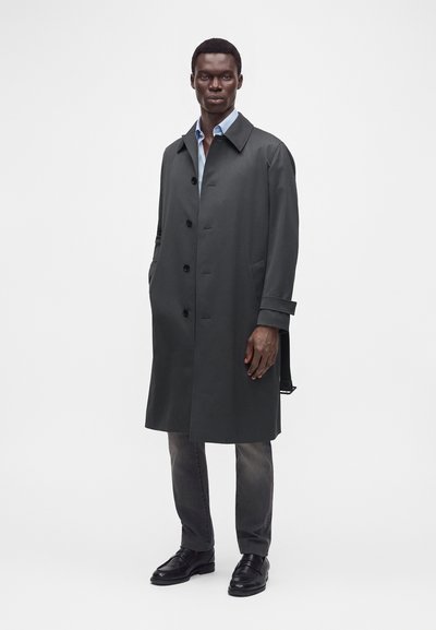 Homme portant un long manteau gris foncé boutonné sur une chemise bleu clair, un pantalon gris et des chaussures en cuir noires sur un fond blanc uni.