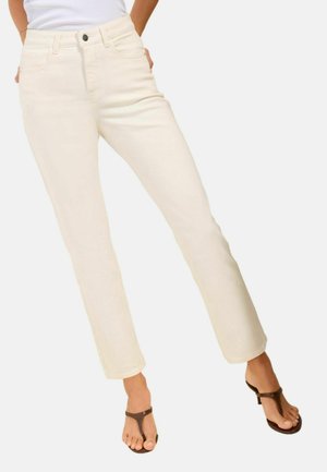 Donna con jeans diritto color avorio e sandali infradito marroni, con le mani riposanti dietro i fianchi, in piedi su uno sfondo chiaro.