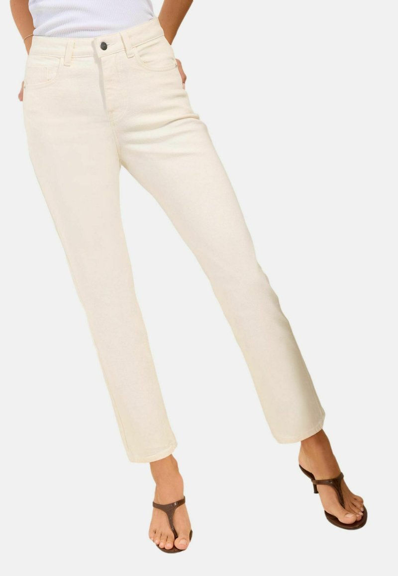 Kvinde iført cremefarvede jeans med lige ben og brune thongsandaler, hænderne hvilende bag hofterne, stående på lys baggrund.