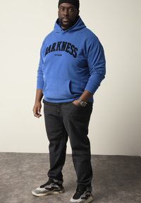 Niebieska bluza z kapturem z czarnym napisem "DARKNESS" i kieszenią kangurką, w połączeniu z ciemnymi dżinsami oraz czarno-szarymi sneakersami.