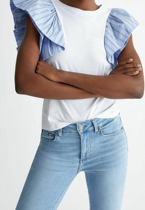 Person krydser arme iført en hvid top med blåstribede, flæseærmer og lyseblå, højtaljede jeans mod en ensfarvet baggrund.