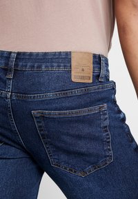 Jeans de mezclilla azul oscuro con un acabado texturizado. Presentan un parche de piel marrón con texto en relieve en la cintura y dos bolsillos traseros.