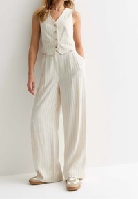 Gilet beige rayé à cinq boutons associé à un pantalon ample à jambes larges. Le tissu a une finition texturée ; baskets avec des accents beiges et blancs.