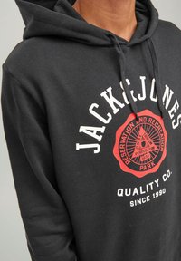Schwarzer Hoodie mit einem Logo, das "JACK & JONES" in Weiß, einem roten runden Abzeichen und dem Text "QUALITY CO. SINCE 1990" zeigt. Aus einem Baumwoll-Mix gefertigt.