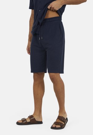 Mężczyzna noszący granatowe szorty ze sznurkiem, ciemnoniebieską koszulę podniesioną, odsłaniającą talię, oraz brązowe sandały z podwójnym paskiem, stojący na białym tle.