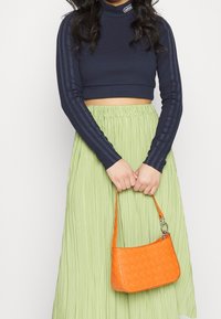 Kvinna klädd i en marinblå, kort Adidas långärmad topp och en ljusgrön veckad kjol, håller en liten kviltad orange handväska.