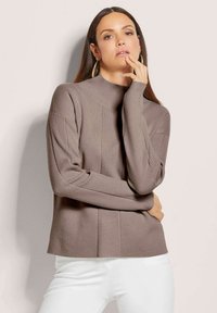 Helles braunes Strickpullover mit hohem Kragen, gerippten Ärmeln und vertikalen Strukturmustern, kombiniert mit weißen Hosen vor einem neutralen Hintergrund.