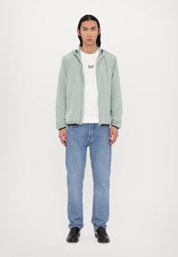 Lys grøn, lynlåset jakke med hætte, båret over en hvid T-shirt og blå jeans. Har en glat overflade og elastiske manchetter. Sorte sko.