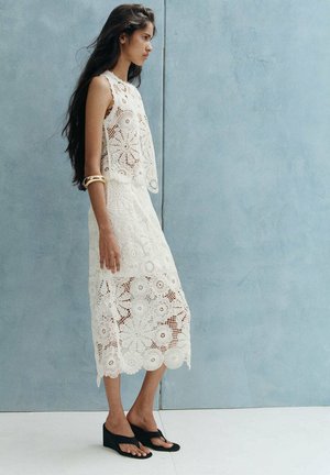 Mujer de pie con un vestido blanco sin mangas de crochet y sandalias negras de cuña frente a una pared azul claro, con pulseras doradas.
