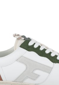 Baskets blanches avec des accents en daim vert, logo gris, tissu texturé et lacets à motif. La semelle est de couleur crème avec des coutures visibles.