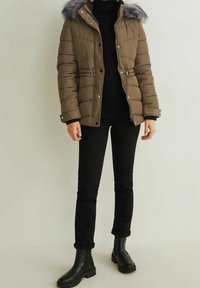 Brown puffer jacket with a fur-trimmed hood, cinched waist, and zippered pockets; worn over a black turtleneck and black pants with ankle boots.